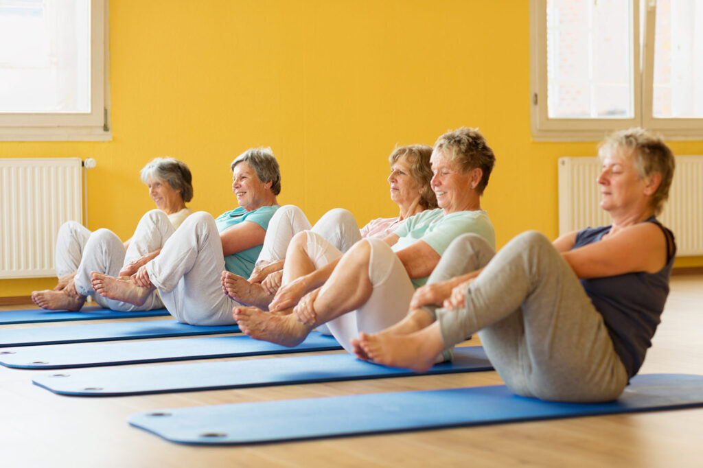 group of active senior women in yoga class exercising on mat in yellow painted room